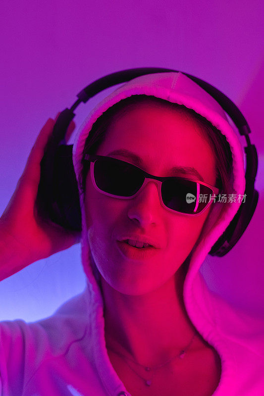 Female portrait in neon light. Fashion woman in purple color. Vibrant background at the studio. Futuristic concept. Girl wear sunglasses and hoodie and listening music in wireless headphones